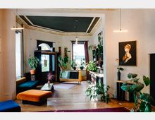 Heller Wohnzimmerbereich mit Holzboden, einer schwarzen Decke mit goldenen Stuckverzierungen und großen Fenstern. Der Raum ist mit Pflanzen, farbenfrohen Sitzmöbeln, Kunstwerken an den Wänden und einem Mix aus modernen und antiken Möbeln eingerichtet.