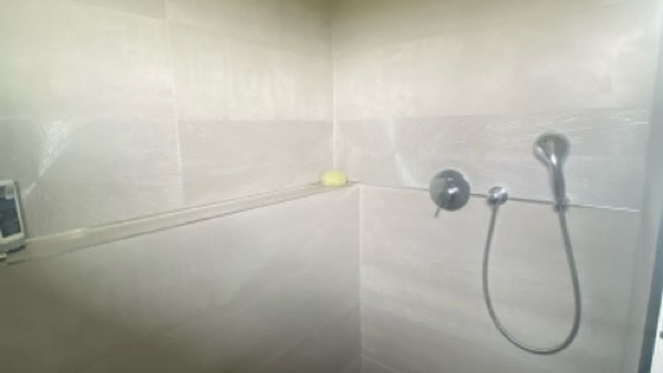 Geflieste Dusche mit hellen Wandfliesen und dunkler Duschwanne, ausgestattet mit großer Regendusche an der Decke und Handbrause an der Wand. In der Ecke verläuft eine Ablageleiste entlang der Wand.