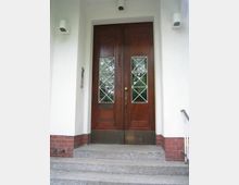 Zweiflügelige Holztür mit Glaseinsätzen, die ein Rautenmuster zeigen, in einer weiß verputzten Gebäudefassade mit roten Ziegeldetails an der Basis. Eine kleine Treppe führt zur Tür, und Überwachungskameras sind an der Wand angebracht.