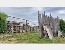 Holzkonstruktion auf einer offenen Wiese, bestehend aus einem einfachen Gerüst aus Baumstämmen und einem teilweise befestigten Tuch. Rechts befindet sich ein hölzerner Zaun, davor liegen Holzplatten und eine Schubkarre, umgeben von niedriger Vegetation und bewölktem Himmel im Hintergrund.
