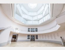 Heller, kreisförmiger Innenraum eines modernen Museums, mit einer Deckenkuppel aus Milchglas, einer gebogenen Treppe und Beschriftungen an den weißen Wänden. Im Hintergrund sind eine Tür, Fenster und ein Ausstellungsraum sichtbar.