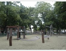 Spielplatz mit Holzkonstruktionen und Seilnetzbrücken, die auf einem sandigen Untergrund stehen. Im Hintergrund sind zahlreiche Bäume sowie Gebäude zu sehen.