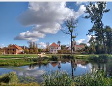 Ländliche Szenerie mit einem Teich im Vordergrund, umgeben von Schilf und grünem Gras; im Hintergrund stehen ein historisches Gebäude mit rotem Ziegeldach, eine helle Kirche mit Turm und ein Glockenturm. Der Himmel ist blau mit wenigen Wolken, und vereinzelt stehen Bäume auf der Wiese.