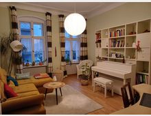 Helles Wohnzimmer mit Holzboden und gelbem Sofa, einem kleinen runden Holztisch, einer weißen Lampe und großen Fenstern mit Vorhängen. Ein weißes Klavier steht neben einem Regal mit Büchern und Dekorationen, und Pflanzen sowie Sitzmöbel ergänzen die Einrichtung.