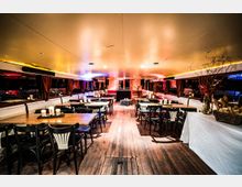 Innenraum eines Veranstaltungsschiffs mit Holzfußboden, gedeckten Tischen und Stühlen. Der Raum ist mit Kerzen beleuchtet, an den Seiten befinden sich Fenster mit Vorhängen, und im hinteren Bereich ist eine kleine Bühne mit DJ-Pult sichtbar.