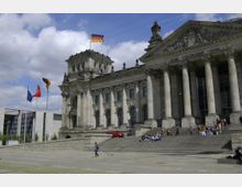 Das Bild zeigt den Reichstag in Berlin, ein historisches Gebäude mit einer neoklassizistischen Fassade, großen Säulen und einer Inschrift „Dem Deutschen Volke“ auf dem Giebel. Vor dem Gebäude befinden sich eine Treppe, mehrere Menschen und wehende Flaggen, darunter die deutsche und die europäische Flagge.