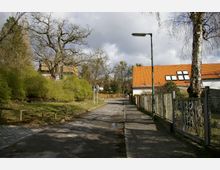 Ländliche Straße mit Gehweg, umgeben von hohen Bäumen und Sträuchern auf der linken Seite sowie einem Zaun und Häusern mit orangefarbenen Ziegeldächern auf der rechten Seite. Bewölkter Himmel und eine einzelne Laterne am Straßenrand.