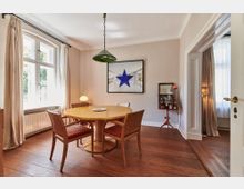 Esszimmer mit Holzboden, einem runden Esstisch aus hellem Holz und vier passenden Stühlen mit roten Polstern. Große Fenster mit beigen Vorhängen lassen Tageslicht herein, und an der Wand hängt ein Bild mit einem blauen Stern.