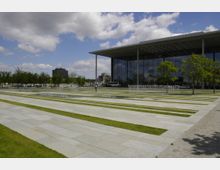 Großer moderner Platz mit rechteckigen Stein- und Rasenstreifen, umgeben von kleinen Bäumen. Im Hintergrund ein Gebäude mit einer verglasten Fassade und einem flachen Dach; im Vordergrund Wasserspiele mit symmetrisch angeordneten Fontänen.