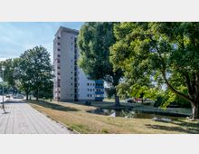 Ein mehrstöckiges Wohngebäude mit Balkonen steht in einer parkähnlichen Umgebung mit Wiesen, großen Bäumen und einem kleinen Wasserbecken. Im Vordergrund führt ein gepflasterter Gehweg an Straßenlampen und parkenden Autos vorbei.