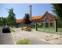 Straßenansicht eines Ziegelgebäudes mit markanten Rundbogenfenstern und rotem Dach, umgeben von einem grünen Zaun. Im Hintergrund befindet sich ein hoher Schornstein, und entlang der Straße parken mehrere Autos.