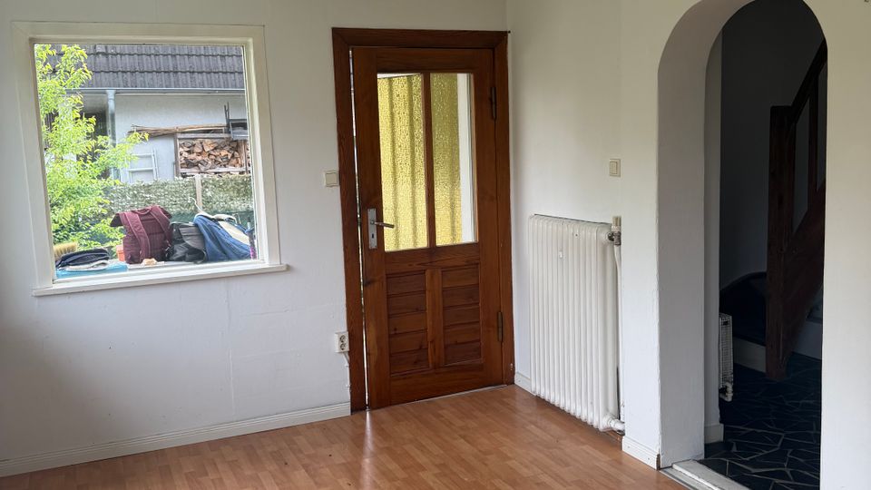 Leerer Wohnraum mit hellem Laminatboden und weißen Wänden, mit einem Fenster zur Straße und einer Holztür mit gelblich getöntem Glaseinsatz. Rechts führt ein Rundbogen-Durchgang in einen angrenzenden Bereich; daneben hängt ein weißer Heizkörper an der Wand.