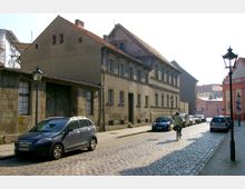 Straßenszene mit Kopfsteinpflaster und einer Reihe von geparkten Autos entlang eines historischen Gebäudes mit verputzter Fassade und Ziegeldach. Im Hintergrund sind weitere Gebäude sowie eine Person auf einem Fahrrad sichtbar.