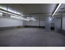 Großer, leerer Raum mit grauem Betonboden und weißen Wänden, teils mit grauer Verkleidung. Die Decke besteht aus Metallpaneelen und ist mit langen, leuchtenden Deckenleuchten ausgestattet; im Hintergrund sind eine Tür und eine Metallwand sichtbar.