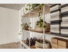 Offenes Regal in einem modernen Innenraum, gefüllt mit Pflanzen, geflochtenen Körben, Glasflaschen und gestapelten Yogablöcken. Der Raum hat eine helle Wand, eine Holzoptik am Boden und eine Betondecke.