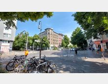 Straßenecke mit mehrstöckigem historischen Gebäude im Zentrum, umgeben von Bäumen und Gehwegen. Im Vordergrund sind parkende Fahrräder an Ständern sichtbar, während Passanten die gepflasterten Gehwege entlanggehen; Geschäfte und Straßenschilder rahmen die Szene ein.
