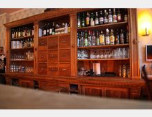 Holztheke in einer Bar mit einem großen Regal, das verschiedene Flaschen von Alkoholika und Weinflaschen enthält. Im unteren Bereich sind Gläser sowie kleine Schubladen und offene Ablagen zu sehen.