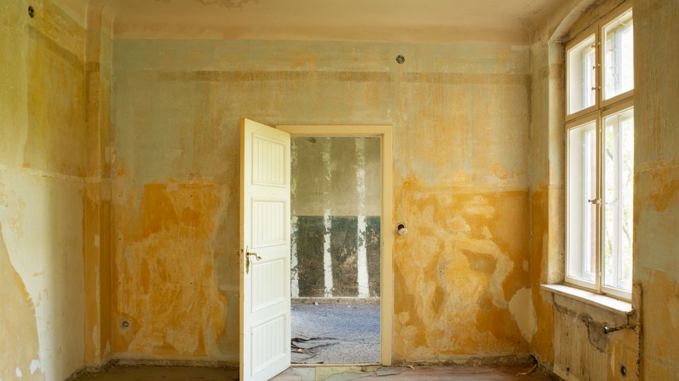 Unrenovierter Innenraum eines alten Gebäudes mit abblätterndem Putz und freigelegten Wandflächen, Holzboden und offener weißer Tür zu einem weiteren kahlen Raum. Rechts sorgt ein großes Sprossenfenster mit tiefer Laibung für Tageslicht.