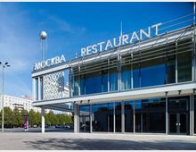 Moderne Glasfassade eines zweistöckigen Gebäudes mit der Aufschrift „Mockba Restaurant” in großen Buchstaben auf dem Dach; im Vordergrund eine freie Fläche und links im Hintergrund Bäume und Wohngebäude.