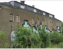 Verlassenes Gebäude mit einer bräunlichen Fassade und mehreren zugemauerten oder mit Brettern verschlossenen Fenstern im Obergeschoss. Die Außenwand ist mit Graffiti bedeckt, und dichter Bewuchs mit Bäumen und Sträuchern umgibt das Gebäude.