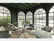 Wintergarten mit einem grünen Holzrahmen und großen Sprossenfenstern, die den Blick auf Schilf und ein Gewässer freigeben. Im Innenraum befinden sich einfache Möbel wie ein Sofa, Stühle und Tische aus Metall sowie Kissen für eine gemütliche Atmosphäre.