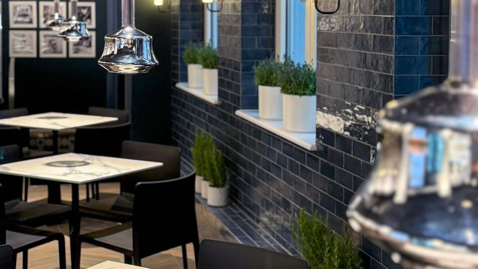 Modern eingerichtetes Café mit Tischen aus weißem Marmorimitat, schwarzen Stühlen und einer Wandverkleidung aus glänzenden dunkelblauen Fliesen. An der Wand befinden sich rechteckige Wandleuchten und Fenster mit kleinen Pflanzen in weißen Töpfen auf den Fensterbänken.