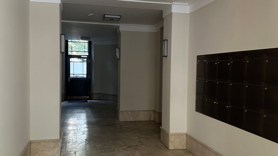 Heller Eingangsflur eines Wohngebäudes mit beigefarbenen Wänden, Steinfliesenboden und abgehängter Decke mit indirekter Beleuchtung. Rechts befindet sich eine Wand mit Briefkastenanlage, am Ende des Flurs eine Glastür mit Blick nach draußen.