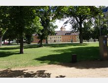 Ein Park mit einer Rasenfläche, umgeben von großen Bäumen. Im Hintergrund steht ein langer, zweigeschossiger Gebäudekomplex in gelben und orangefarbenen Fassaden, mit einem Kirchturm in der Mitte.