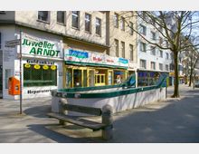 Straßenszene mit mehreren Geschäften in einem mehrstöckigen Gebäude, darunter ein Juweliergeschäft, ein Reformhaus und andere Geschäfte. Im Vordergrund befinden sich ein Holzsitzbank, ein Betonpflanzkübel und der Zugang zu einer U-Bahn-Station mit grünem Geländer.