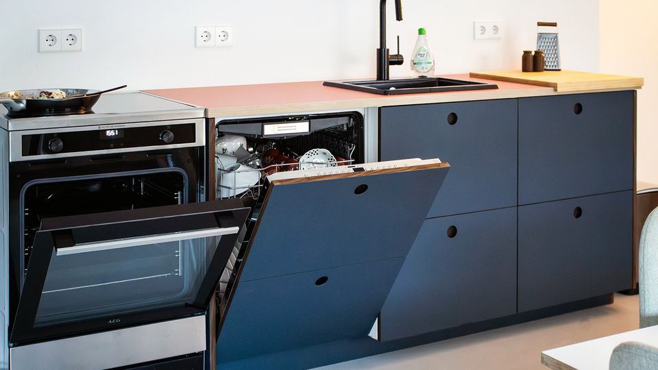 Moderne Küchenzeile mit dunklen, grifflochgefrästen Fronten und Holzarbeitsplatte, ausgestattet mit Einbaubackofen, schwarzer Spüle und hoher Armatur. Die Spülmaschine ist geöffnet und zeigt den Geschirrkorb; an der weißen Wand sind mehrere Steckdosen zu sehen.