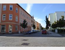 Straßenecke mit gepflasterten Gehwegen, mehrstöckigen Wohngebäuden mit bunten Fassaden in Orange und Grün, geparkten Autos entlang der Straße sowie vereinzelten Bäumen am Gehwegrand. Der Himmel ist klar und blau.