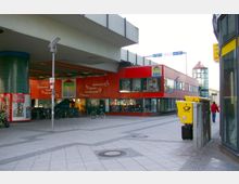 Ein moderner Platz mit einer roten, zweistöckigen Gebäudefassade, die Geschäfte und Schaufenster zeigt, sowie einem überhängenden Brückenteil darüber. Im Vordergrund stehen gelbe Briefkästen und Fahrräder, während im Hintergrund Glasfassaden und Gehwege erkennbar sind.