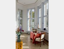 Heller Wintergarten- oder Erkerraum mit hohen, weiß gerahmten Sprossenfenstern an zwei Seiten und hellem Steinboden. In der Mitte steht ein kleiner Tisch mit roter Tischdecke und Kerzen, daneben ein gepolsterter Holzstuhl; entlang der Fenster verläuft ein Heizkörper.