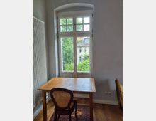 Holzgetäfeltes Zimmer mit einem rechteckigen Holztisch und einem Stuhl mit geflochtener Lehne. Im Hintergrund ein großes, weißes Doppelfenster mit Sprossen, das den Blick auf grüne Bäume und ein Gebäude mit rotem Dach freigibt.