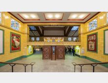 Historische Bahnhofshalle mit gelb gefliesten Wänden und dekorativen Holzelementen, einschließlich einer großen Holztür im Zentrum. An den Wänden hängen retroartige Werbeplakate, und im Hintergrund sind ein Fahrkartenautomat und eine Menschenmenge zu sehen.