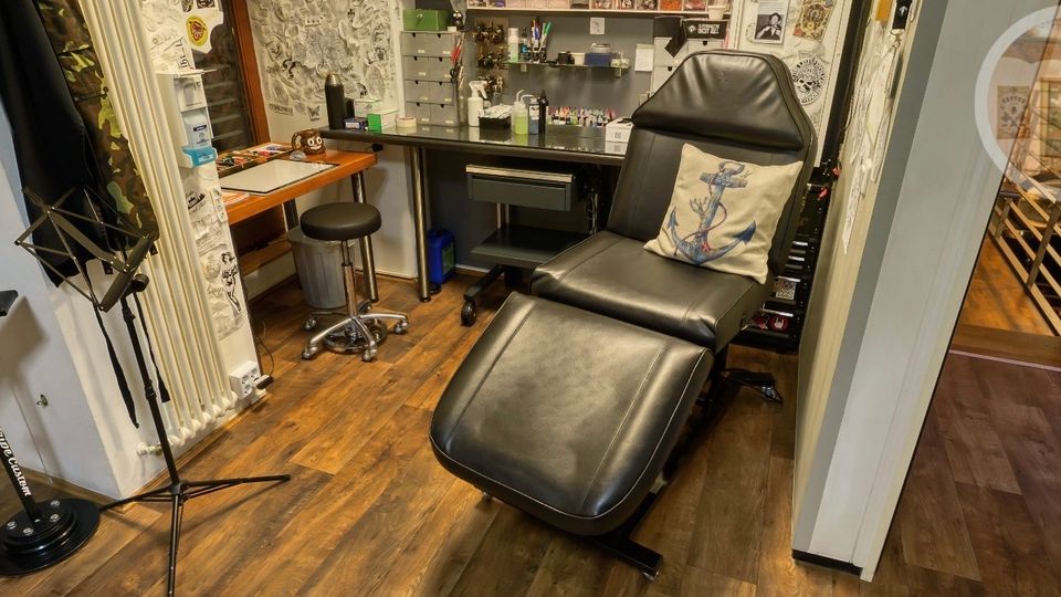 Kleines Tattoo-Studio mit schwarzer Behandlungsliege im Vordergrund, Holzfußboden und Wänden voller Zeichnungen und gerahmter Kunst. Im Hintergrund stehen ein Arbeitstisch mit Hockern sowie Regale und Schubladen mit Tattoo-Utensilien und Lampen.