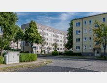 Mehrstöckige Wohngebäude in einer Wohnsiedlung, umgeben von Gehwegen, Bäumen und gepflegten Hecken. Im Vordergrund ist eine gepflasterte Straße und ein Stromverteilerkasten zu sehen.