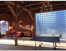 Ein rustikaler Raum mit sichtbaren Holzbalken und einer Ziegelwand, ergänzt durch eine große Glasbausteinwand, die Licht hereinlässt. Im Raum befinden sich ein Tischtennistisch, ein rotes Sofa, eine Stehlampe und weitere dekorative Elemente.