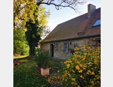 Backsteinhaus mit rotem Satteldach in einer herbstlichen Gartenszene. Im Vordergrund stehen gelb belaubte Büsche und Pflanzkübel, während der Garten mit herabgefallenen Blättern bedeckt ist; ein Baum und ein Kinderfahrzeug befinden sich links.