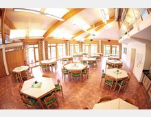 Ein heller Speisesaal mit Holzbalkendecke, großen Fenstern mit bunten Vorhängen und hexagonalen Tischen, die von Holzstühlen mit grünen Polstern umgeben sind. Der Raum hat einen terrakottafarbenen Fliesenboden und ist großzügig mit Tageslicht beleuchtet.