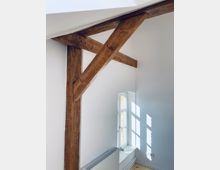 Innenansicht eines Raumes mit sichtbaren Holzträgern an der Wand und der Decke, einem schmalen, weiß gerahmten Fenster, durch das Sonnenlicht fällt, sowie einer weißen Heizung unterhalb des Fensters. Die Wände sind weiß gestrichen und der Boden besteht aus hellem Holz.