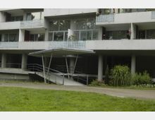 Mehrstöckiges Wohngebäude mit modernen, weißen Balkonen aus perforiertem Metall und großen Glasfenstern. Im Vordergrund eine überdachte Rampe mit Geländern, umgeben von einer Grünfläche mit Pflanzen und Blumen.