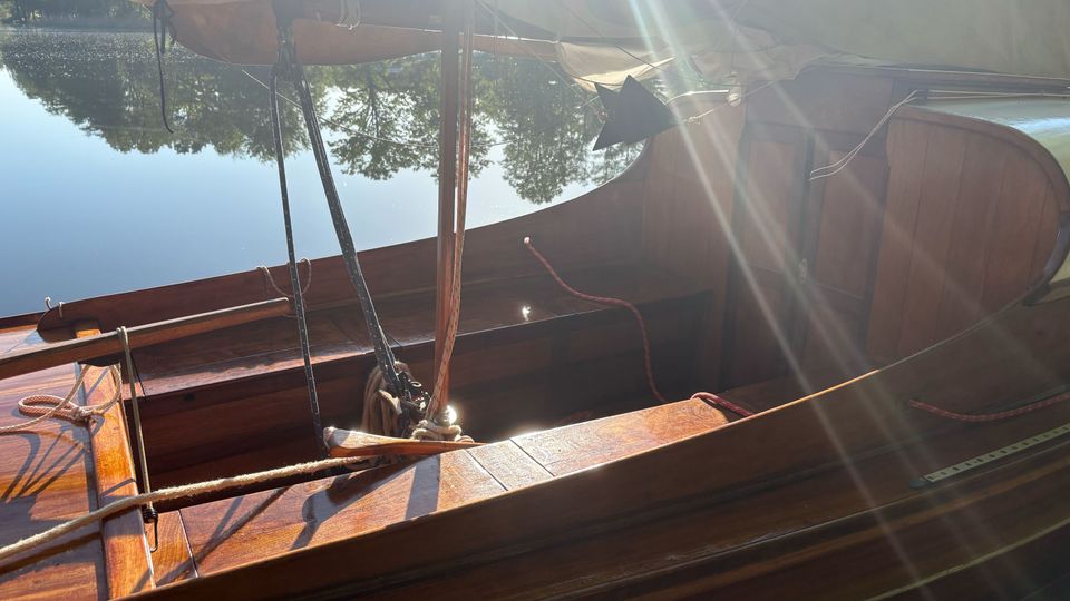 Blick ins Innere eines hölzernen Segelboots mit Mast, Tauwerk und aufgerolltem Segel, das auf einem ruhigen See liegt. Im Hintergrund spiegeln sich Bäume am Ufer im Wasser, während Sonnenstrahlen über das Boot fallen.