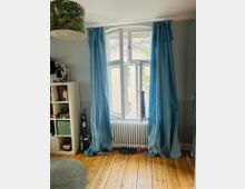 Ein Zimmer mit einem großen, weißen Sprossenfenster, das von blauen Vorhängen eingerahmt ist. Vor dem Fenster steht ein Heizkörper, darunter liegen Schuhe auf einem Holzfußboden; links ist ein Regal mit Spielzeug und Dekorationen zu sehen.