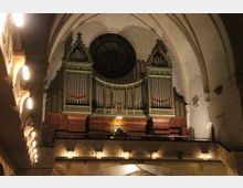 Innenraum einer Kirche mit Blick auf eine große historische Orgel, die auf einer Empore unter einem gotischen Gewölbe steht. Die Orgel hat reich verzierte Pfeifen und wird von einem runden Fenster im Hintergrund überragt, während Wandleuchten die Szene beleuchten.