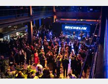 Eine Veranstaltungshalle mit einer großen Menschenmenge, die an Stehtischen und in Gesprächsgruppen verteilt ist. Im Hintergrund befindet sich eine Bühne mit Live-Musikern, während das Wort "Michelin" leuchtend über der Bühne angezeigt wird.