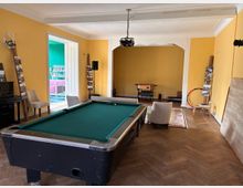 Großer Innenraum mit gelb gestrichenen Wänden, Holzboden und einem Billardtisch im Vordergrund. Hinten befindet sich eine breite Durchgangsöffnung mit einem hängenden Deckenprojektor, daneben Stühle, Regale und eine offene Tür zu einem angrenzenden, farbig gestrichenen Raum.