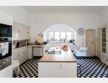 Helle Küche mit einem schwarz-weiß karierten Boden, weißen Schränken und einer Kücheninsel mit einer Holzarbeitsplatte. Im Hintergrund befindet sich ein großes Fenster mit Pflanzen und eine kleine Spüle, die von weißen Fliesen umgeben ist.