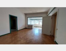 Wohnraum mit Parkettboden, hellen Wänden und grünen Tür- und Fensterrahmen. Im Hintergrund befindet sich ein großer, heller Raum mit mehreren Fenstern, die Tageslicht hereinlassen; ein Heizkörper steht vor einer Innenwandöffnung.