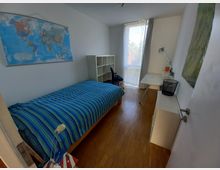 Ein schlichtes Schlafzimmer mit Holzboden, einem Einzelbett mit gestreiftem blau-grünem Bettbezug, einem Schreibtisch mit Lampe, einem Regal und einem großen Fenster mit halbtransparenten Vorhängen. An der Wand hängt eine Weltkarte.